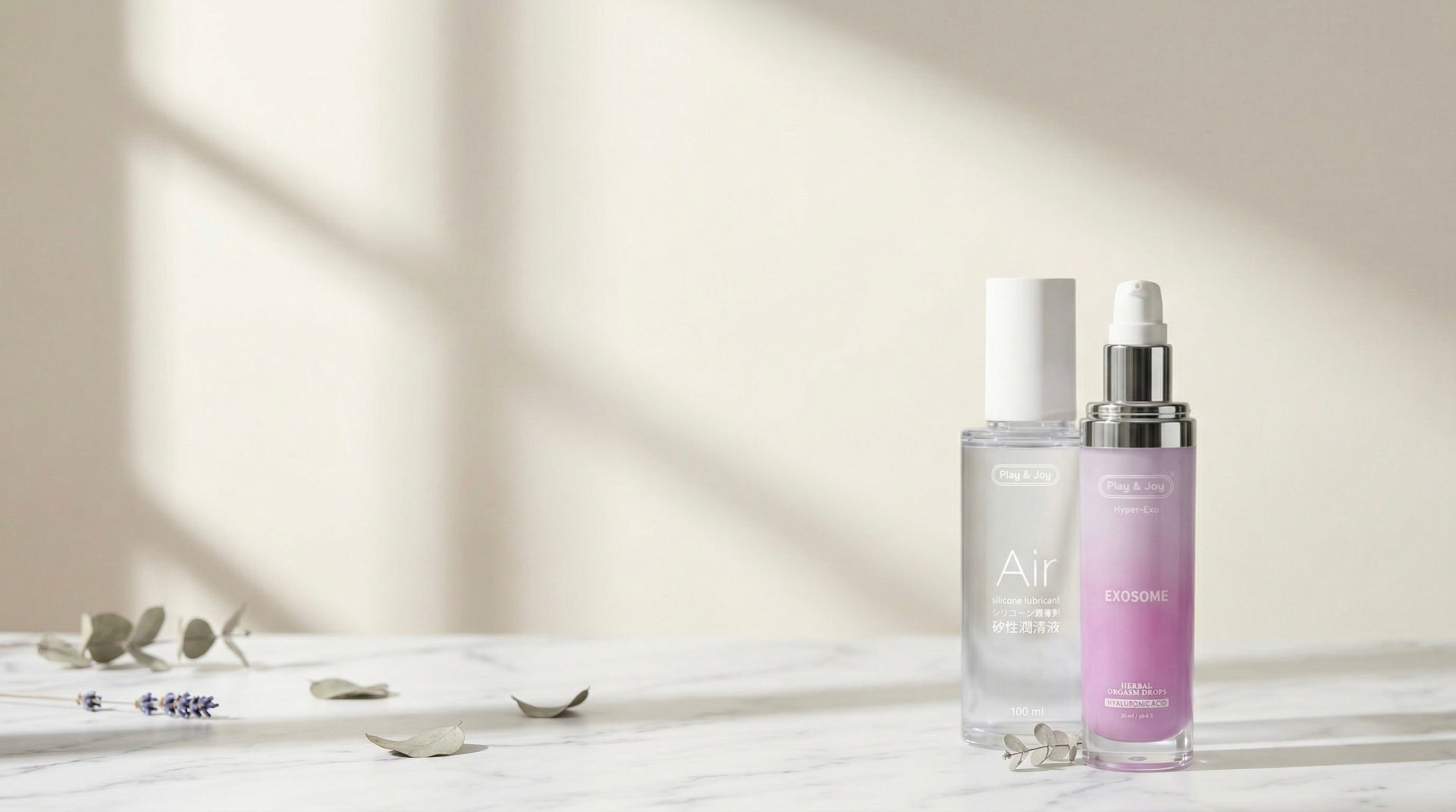 Lubricante de silicona Air y sérum de exosomas Hyper-Exo de Play&Joy Clinical sobre superficie de mármol blanco con ramas de eucalipto y lavanda, dermocosmética íntima de grado médico.